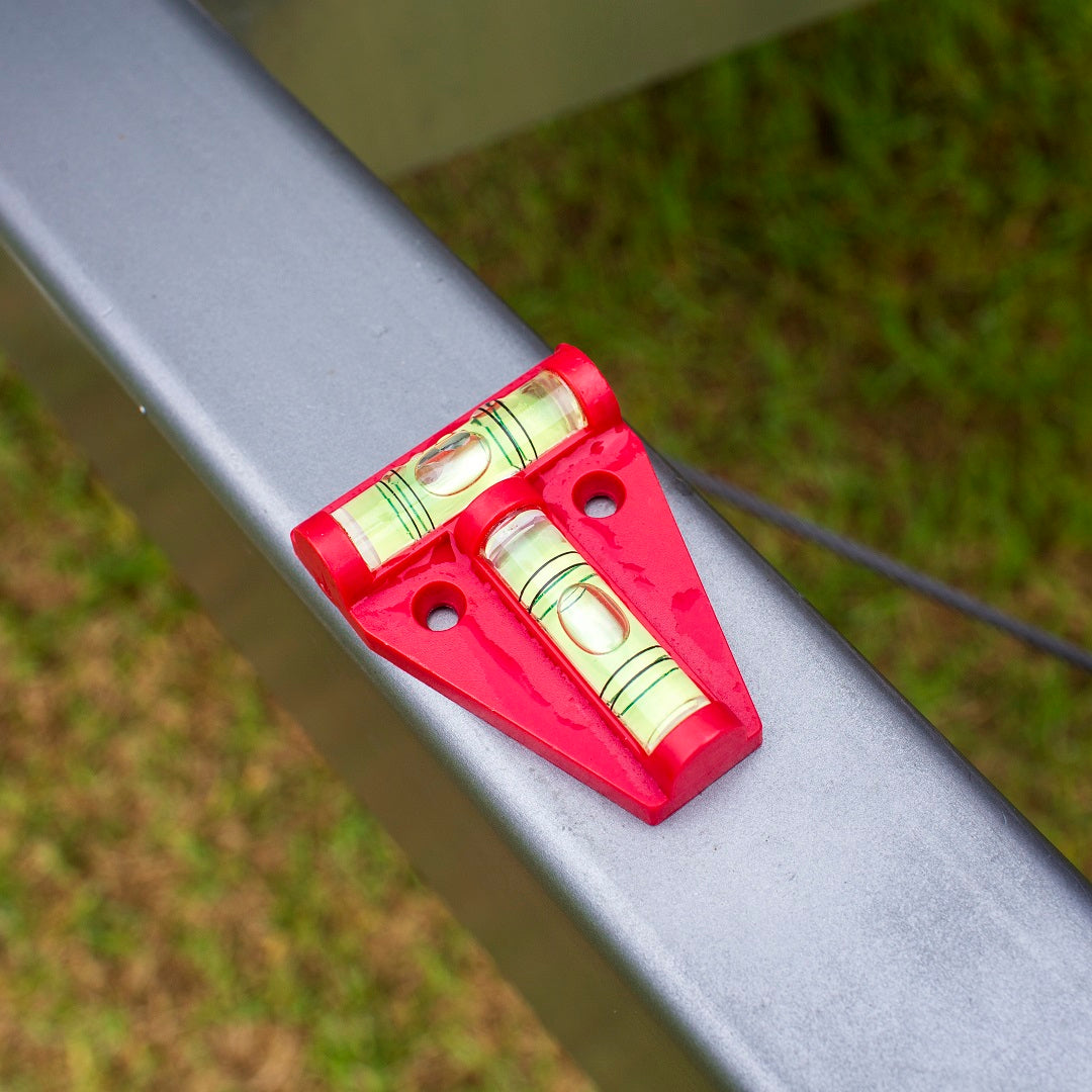 The Explore Caravan T Bubble Level sits on a metal beam with its horizontal and vertical bubbles perfectly centered, indicating a level surface. In the background, grass peeks through, adding a natural touch to this precise moment of RV leveling.