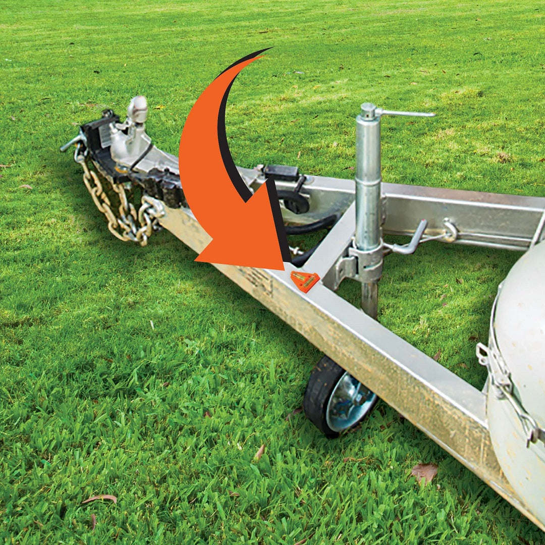 A trailer hitch on green grass shows a red arrow pointing to a small, triangular sticker. Chains, a wheel, and an Explore Caravan T Bubble Level are visible.