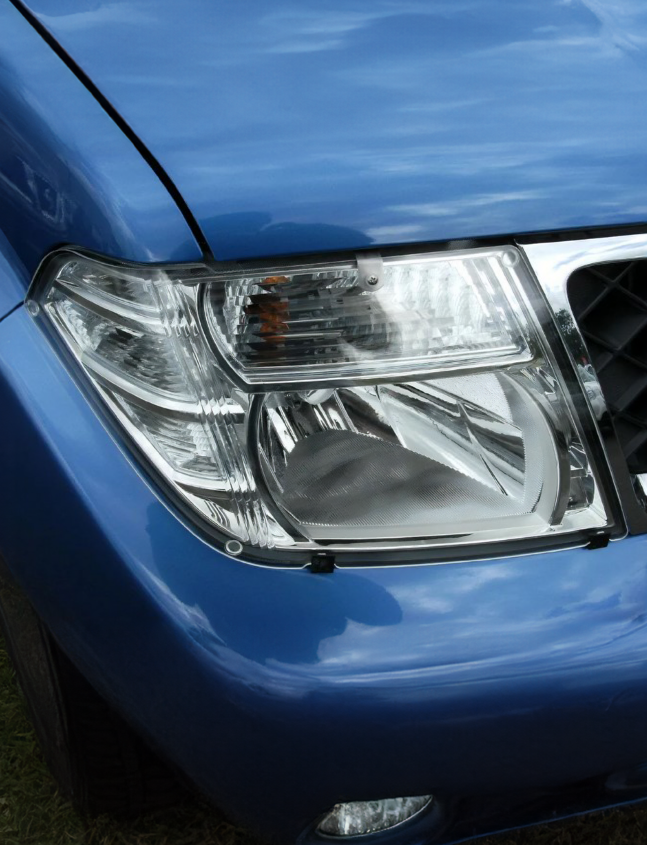 Close-up view of the front right corner of a blue car, featuring a shiny headlight enhanced with Sunland Protective Plastics Headlight Protectors made from UV stable acrylic. The car's hood is visible under a partly cloudy sky, showcasing its custom-moulded chrome grille.