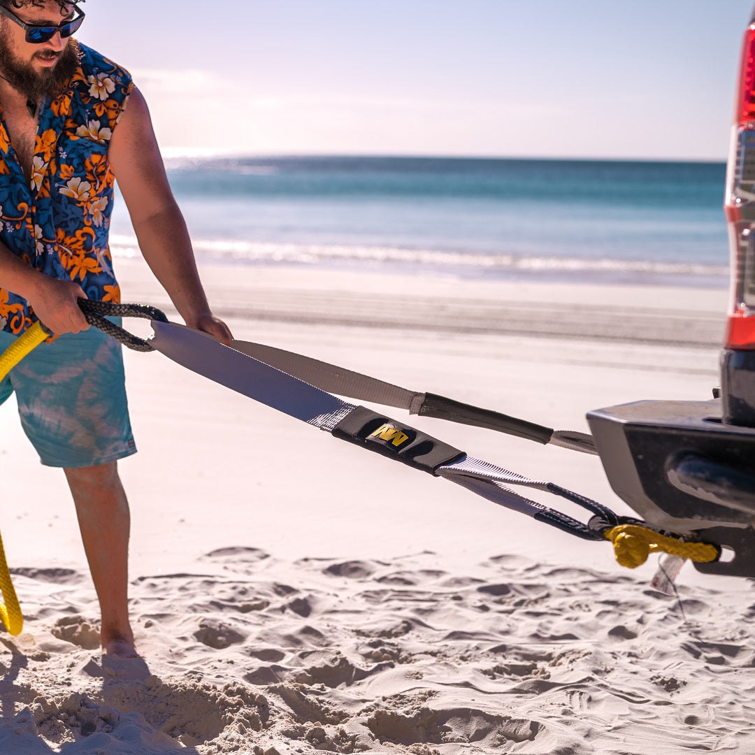 A bearded man wearing sunglasses and a vibrant floral shirt is attaching a Mean Mother Equaliser Strap (75mm / 2.5m, 8,000kg) to the back of a vehicle on a sandy beach. The clear blue ocean and bright sky are visible in the background, indicating he's in the midst of a recovery operation with Mean Mother 4x4 gear.