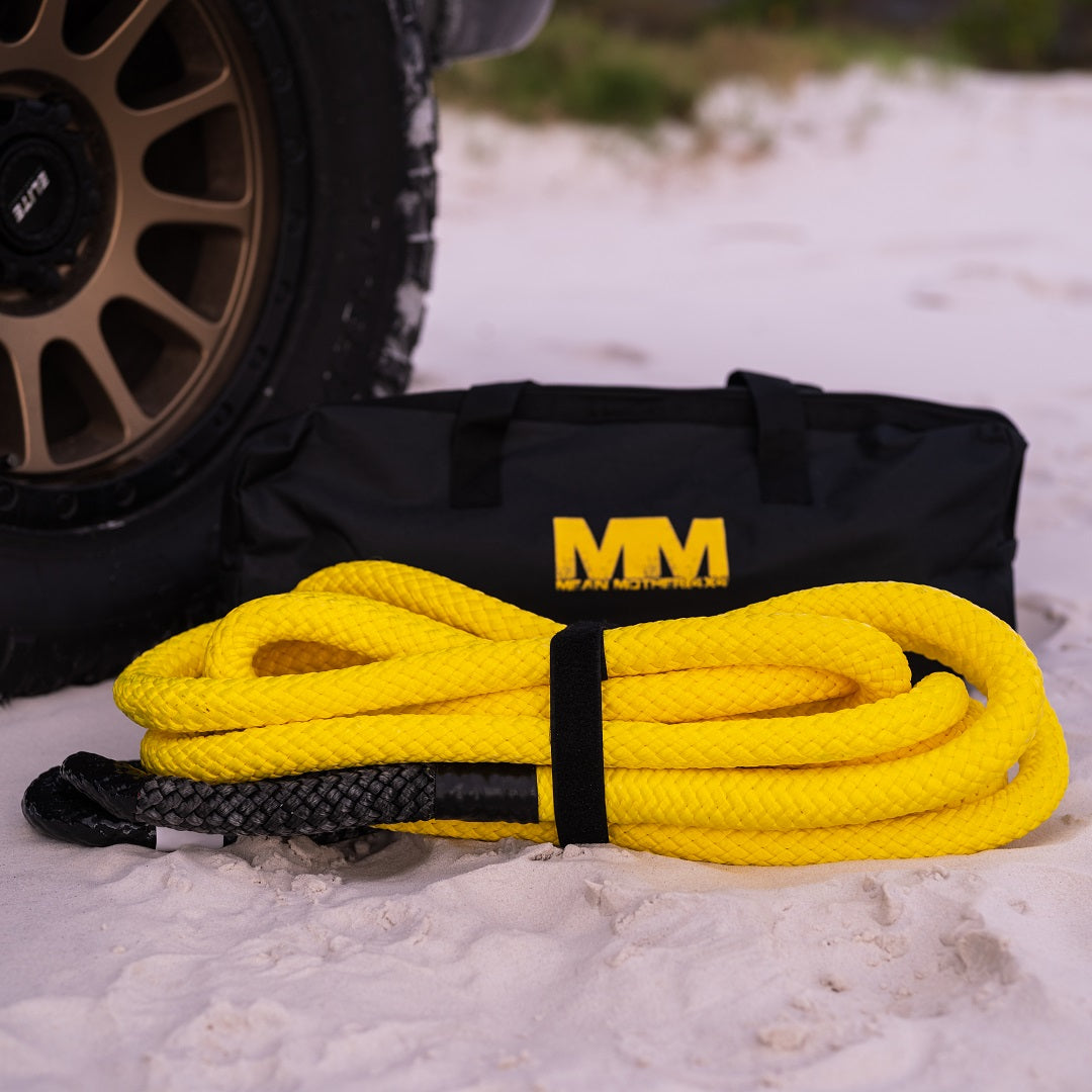 A coiled Mean Mother Kinetic Snatch Rope 9m - 13000kg, noticeable for its vibrant yellow color with black accents and its UV and abrasion resistance, lies on sandy terrain in front of a black bag featuring the yellow initials "MM." A vehicle tire can be seen partially in the background.