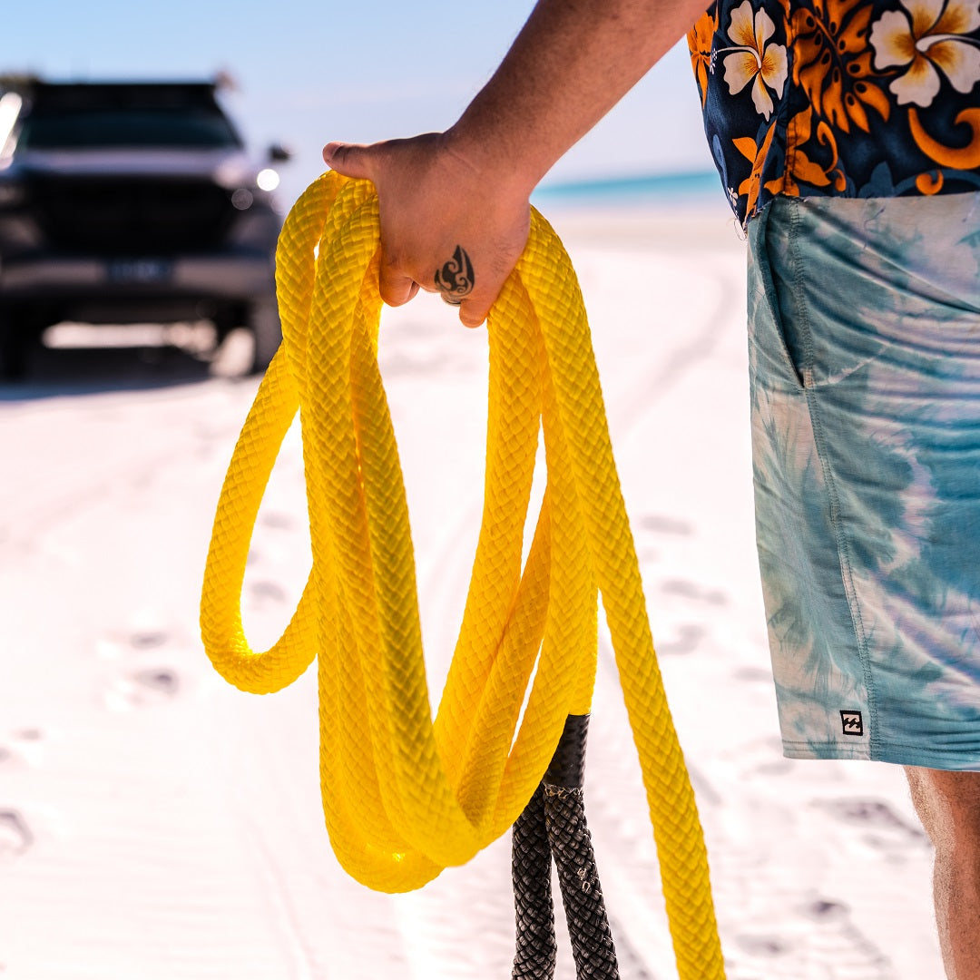 A person on a sandy beach holds the bright yellow Mean Mother Kinetic Snatch Rope 9m - 13000kg, celebrated for its UV and abrasion resistance. They are wearing a floral shirt and blue tie-dye shorts, while a vehicle with an impressive 13000kg Minimum Breaking Strength is parked in the background near the shoreline.