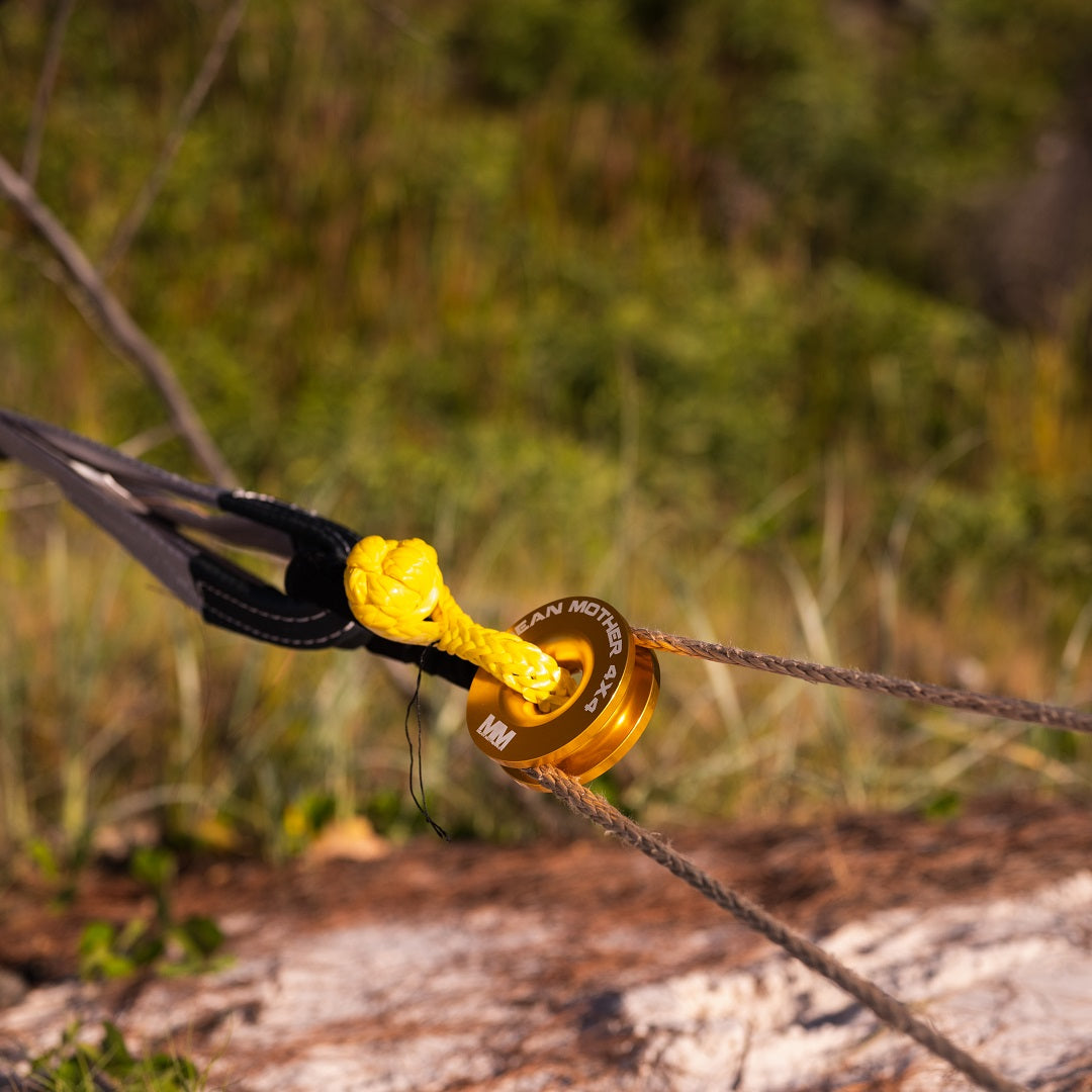 Close-up of a pulley system with a yellow tie on a UHMPE synthetic rope secured to a tree trunk. The pulley displays "Mean Mother 4x4." In the background, blurred greenery and logs suggest an essential component of any robust Soft Shackle & Snatch Ring Bundle for 4x4 recovery.