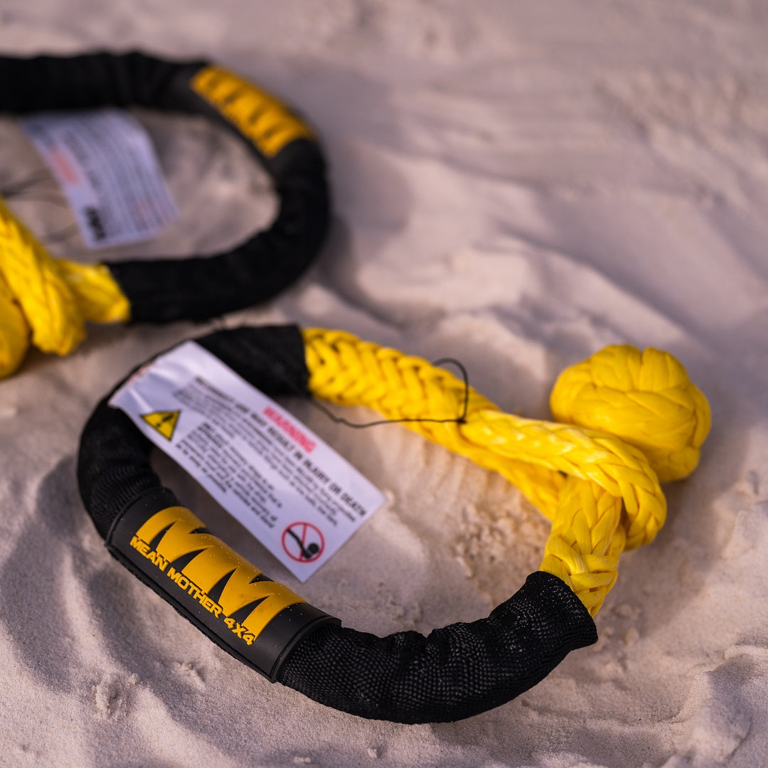 A Soft Shackle 14,700kg from the Mean Mother 4x4 brand, featuring a tightly braided yellow and black design with a protective sleeve and label, rests on a sandy surface, hinting at its inclusion in a recovery kit.