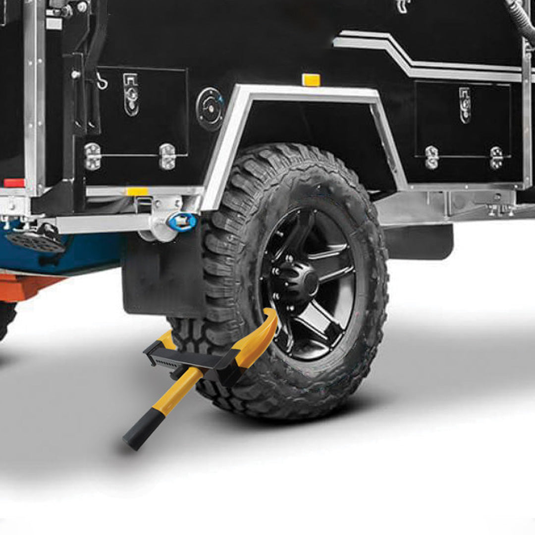 A black tire on a rugged trailer features the "Drive Anti Theft Wheel Clamps" with a yellow ax resting against it, showcasing the clamp's heavy-duty construction. The trailer's body is black with metal accents, set against a white background to highlight its emphasis on caravan security.