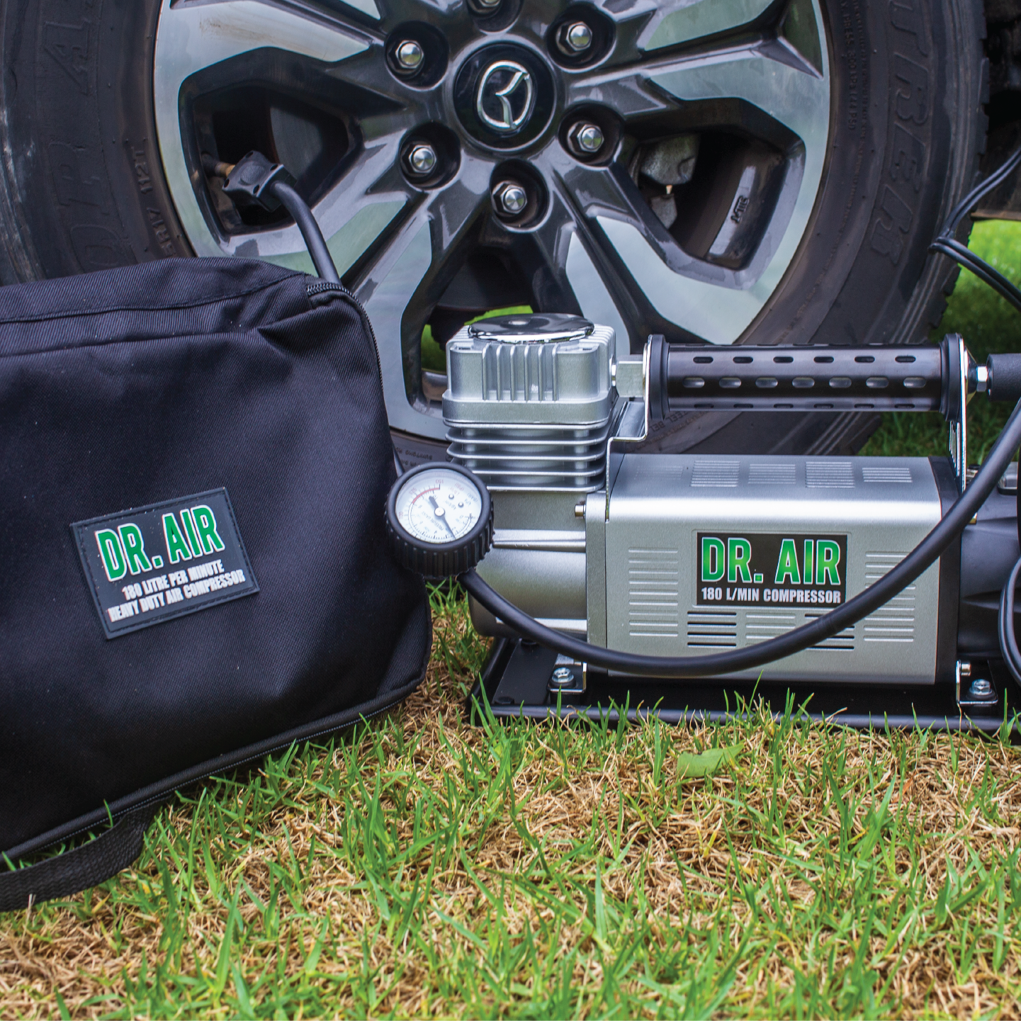 Resting on the grass in front of a car tire is the "Dr Air 12V Air Compressor 180Lpm," offering efficient air delivery with elegance. Its accompanying sleek black carrying bag also bears the "Dr Air" brand. Benefit from a 5-year warranty, ensuring peace of mind as your tire shines with a stylized wheel design.