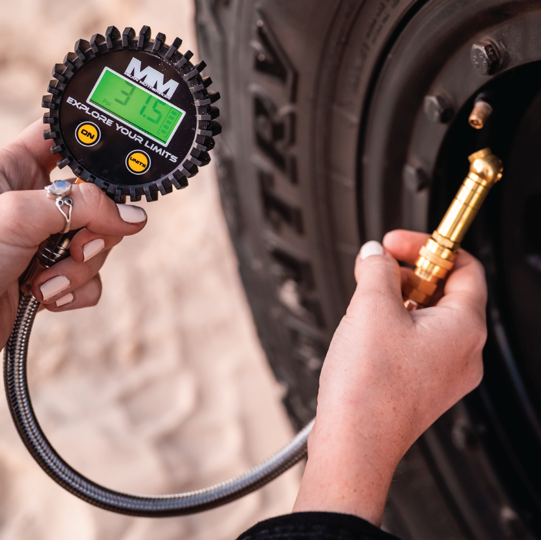 Two hands are shown checking a tire's air pressure using the Mean Mother Digital 2-IN-1 Deflator & Gauge Kit. The gauge displays a reading of 34.5 PSI. The background includes a close-up of the tire tread. The tire's air valve is connected to the digital tyre gauge by a metal hose.

