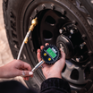 Using the Mean Mother 4x4 Digital Tyre Deflator & Gauge Kit, a person checks the off-road vehicle's tire pressure, with “34.5” displayed for beach driving. The large-tread tire connects to a hose that precisely adjusts the valve.