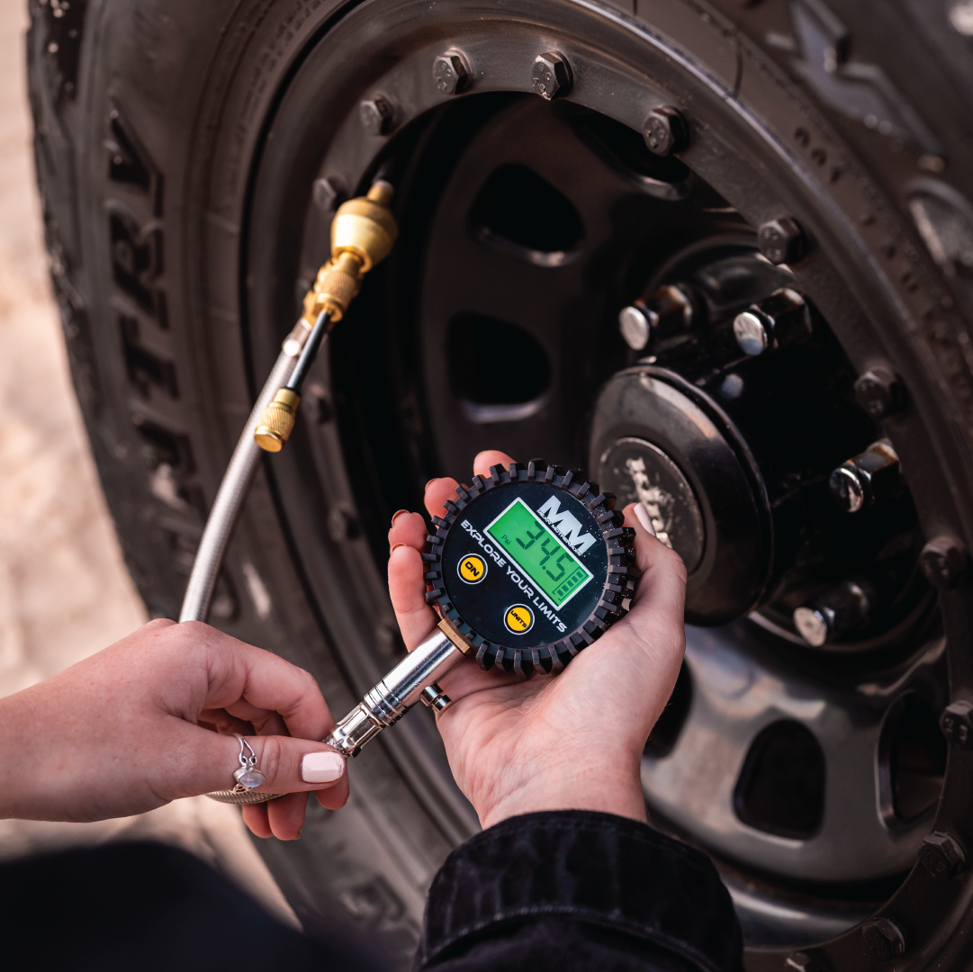 A person uses the Mean Mother Digital 2-IN-1 Deflator & Gauge Kit to measure the pressure of a vehicle's tire, which reads 34.5 PSI. The tire and surrounding equipment are plainly visible, and the person wears a black jacket and has a ring on one of their fingers.