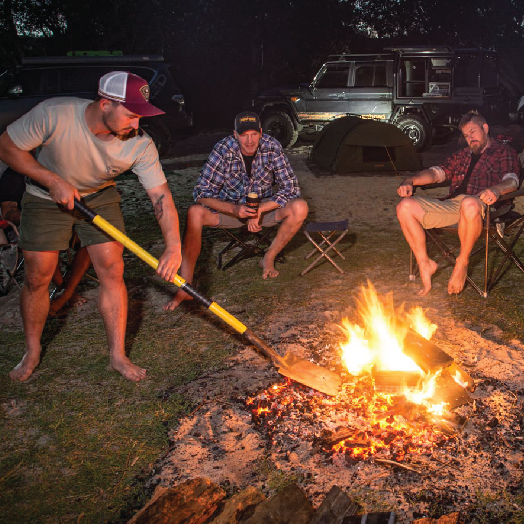 Three men are camping outdoors at night. One, a 4WD enthusiast, is standing and using the Mean Mother 5-IN-1 Multi 4x4 Recovery Shovel (Trade Show Only) to manage the campfire, while the other two sit nearby in camping chairs. They are surrounded by camping gear, including vehicles with Mean Mother 4x4 recovery kits, tents, and coolers. The fire illuminates the scene.