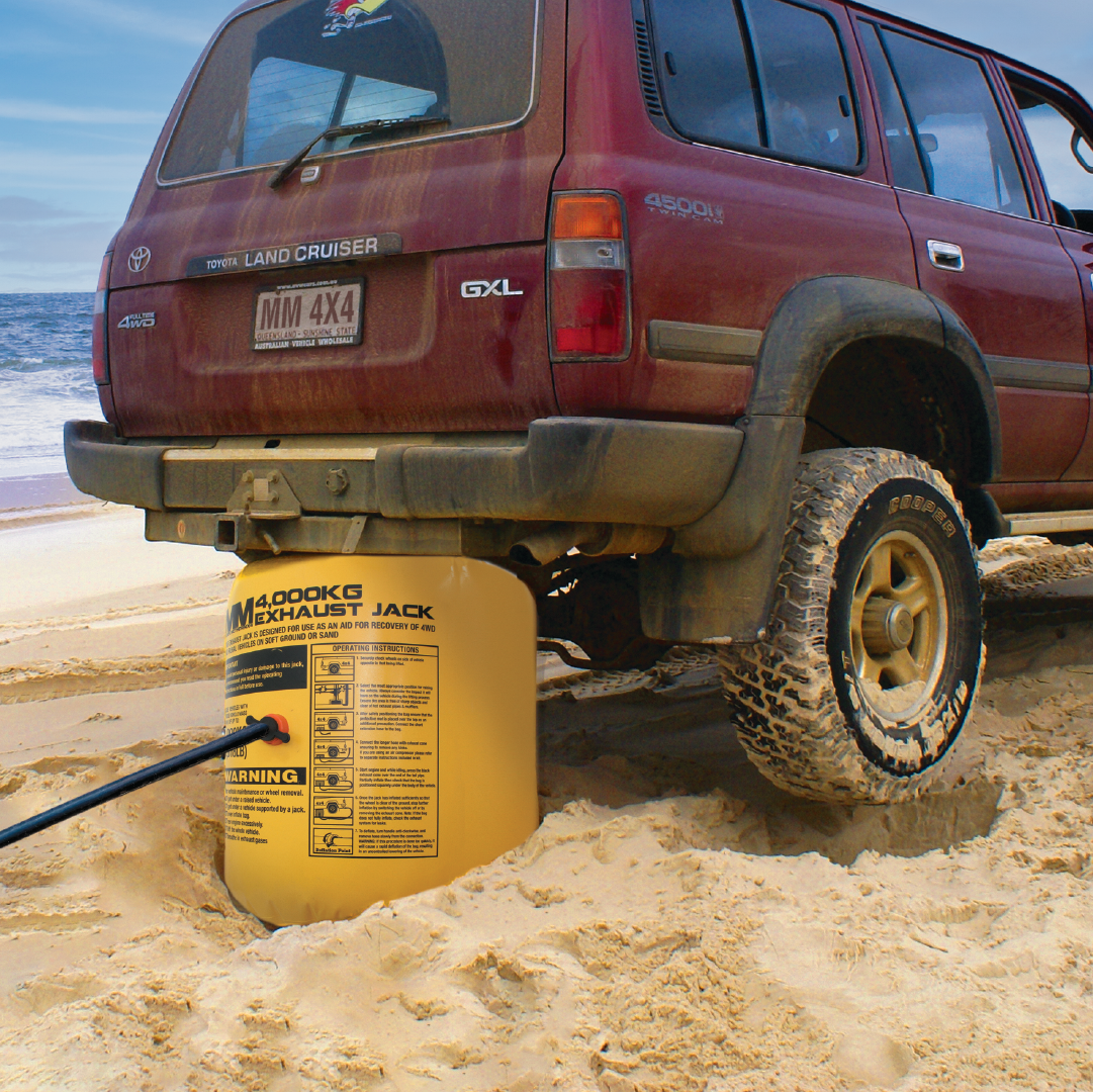 A red Toyota Land Cruiser is stuck in the sand on a beach. A Mean Mother Exhaust / Air Jack 4000kg is placed under the vehicle, lifting it as it inflates. The ocean waves are visible in the background. The exhaust jack is yellow with instructional text and warnings printed on it, perfect for off-road recovery situations by Mean Mother.
