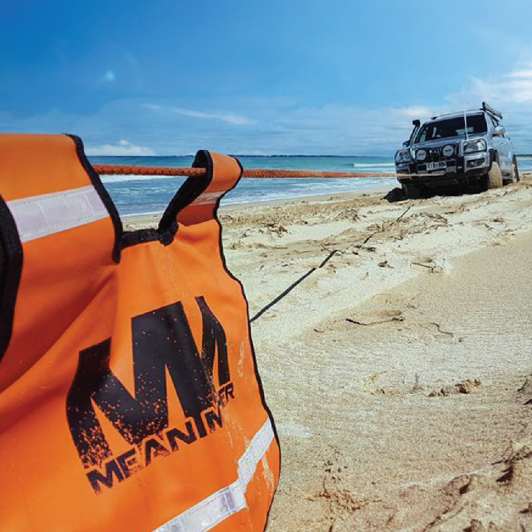 An orange recovery strap extends across the sand towards a 4x4 vehicle stuck on the beach. A high-visibility vest with "Mean-MN" branding and the Mean Mother Recovery Dampener Blanket are positioned in the foreground for safety under the clear blue sky by the ocean.