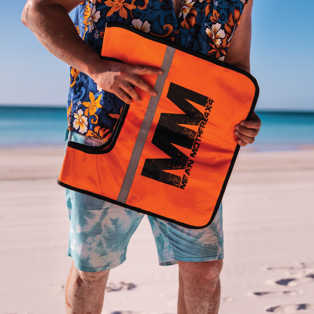 A person wearing a vibrant floral shirt and tie-dye shorts holds an orange bag labeled "Mean Mother," containing the Mean Mother Recovery Dampener Blanket. The backdrop displays a sandy beach and blue ocean under clear skies, creating an ideal blend of relaxation and safety.