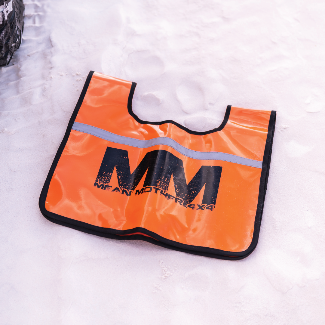 A black and orange Mean Mother Recovery Dampener Blanket, featuring the "MM MEAN MOTHER 4X4" logo, is displayed on a white, snowy surface with a partial view of a textured tire visible to the left.