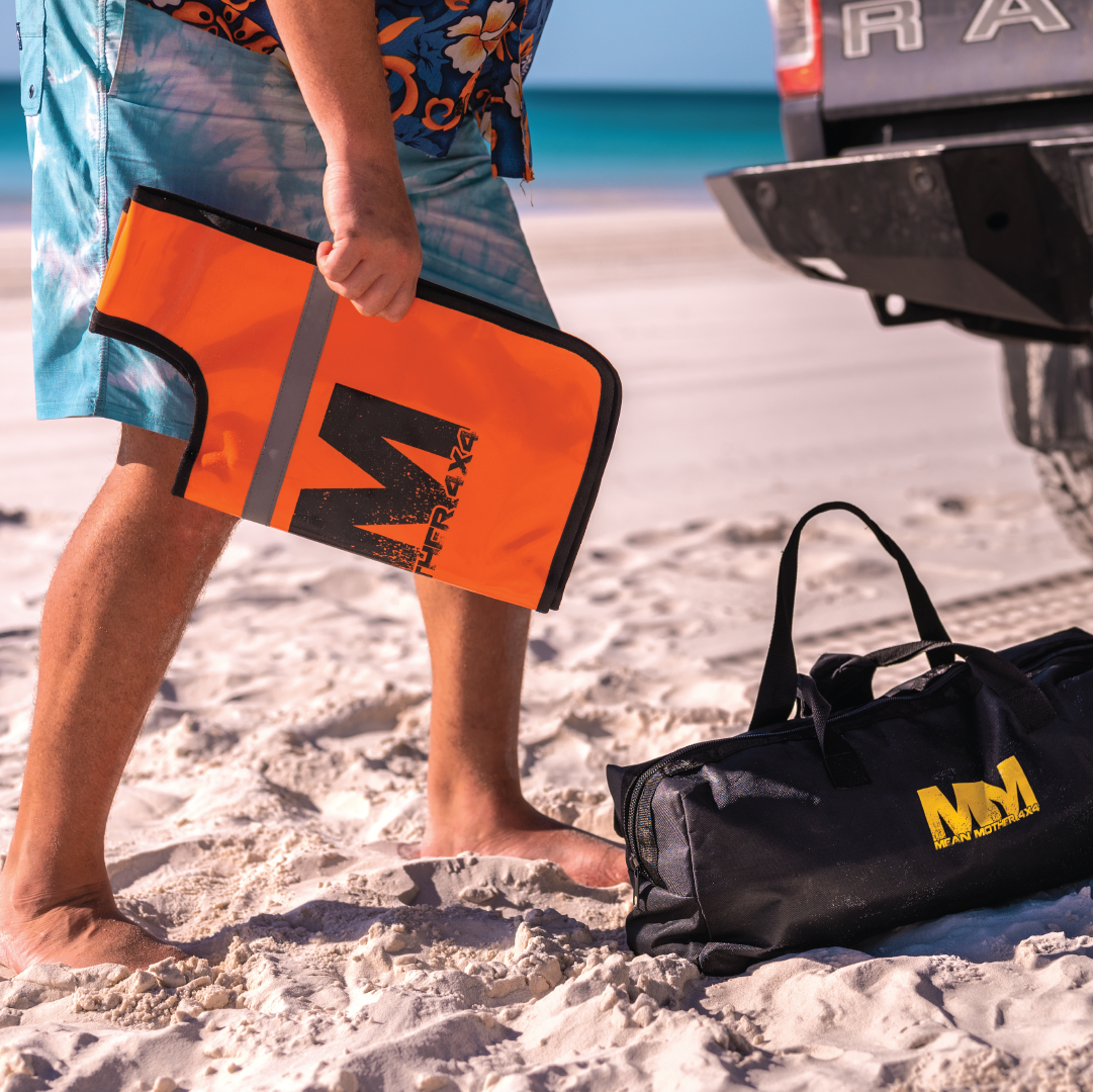 On a sunny beach, a person is holding the Mean Mother Recovery Dampener Blanket in bright orange, featuring the prominent "M" logo, an essential safety accessory for off-road escapades. Nearby on the sand is a black bag adorned with the same logo. A vehicle is parked close by, with turquoise waters shimmering in the background.