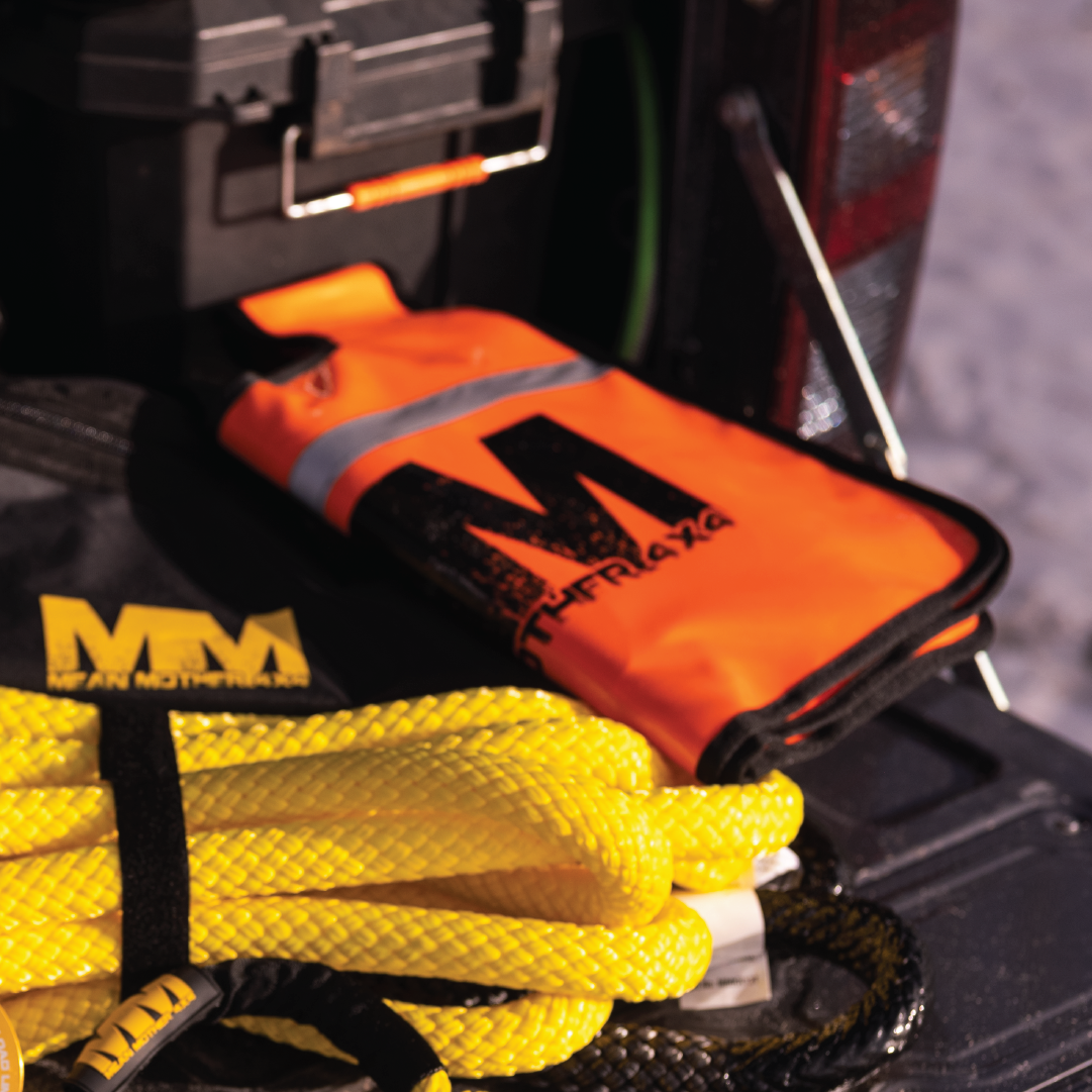 In the truck bed, a Mean Mother Recovery Dampener Blanket in orange with a reflective stripe, a yellow tow rope, and a black toolbox are illuminated by sunlight, casting shadows on these safety accessories.