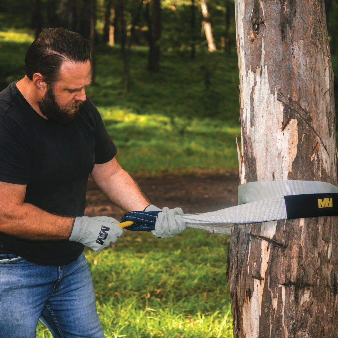 A man in a black shirt and jeans is using a Mean Mother Tree Trunk Protector, which features a blue and yellow polyester webbing, to winch a tree in the forest. He is wearing gray gloves, standing on grass with sunlight filtering through the trees, ensuring the trunk remains unharmed.