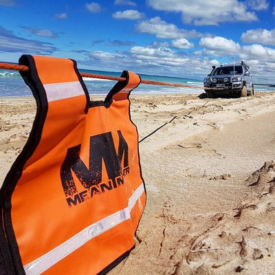 A brightly colored orange recovery damper, emblazoned with "Mean Mother 4x4," is securely attached to a winch cable that stretches across the sandy terrain. In the background, a partially buried 4x4 vehicle is visible near the ocean shore under a blue sky with scattered clouds, ready to utilize its Mean Mother 5pc Recovery Kit 11,000kg Combo Pack and heavy-duty bow shackles for recovery.