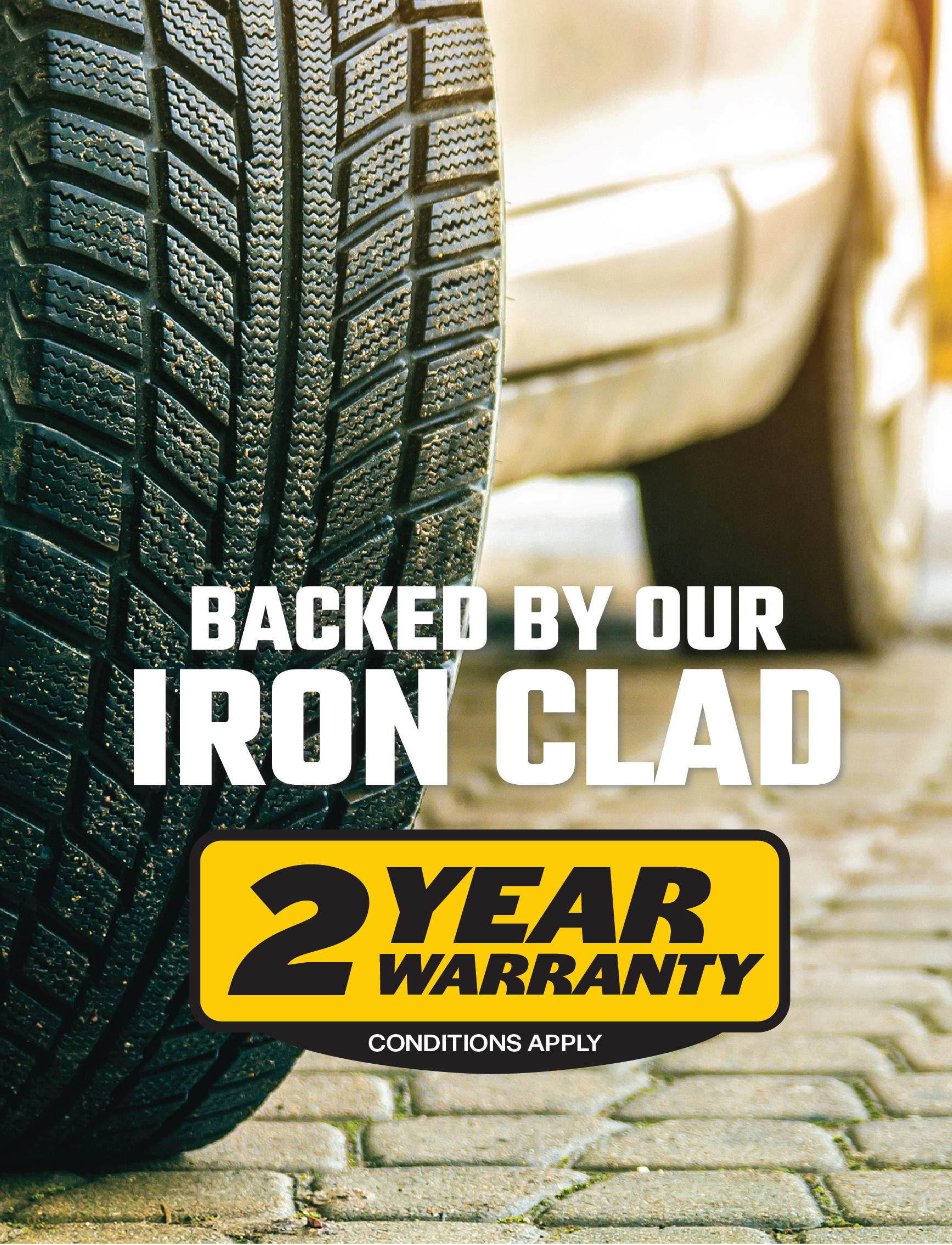 Close-up shot of a car tire on a cobblestone surface with text overlay reading "BACKED BY OUR IRON CLAD 2 YEAR WARRANTY CONDITIONS APPLY." The yellow and black label, similar to the one on our Sunland-Protection Battery Charger 12v 2.8Amp with Overload Protection, emphasizes the two-year warranty offer.